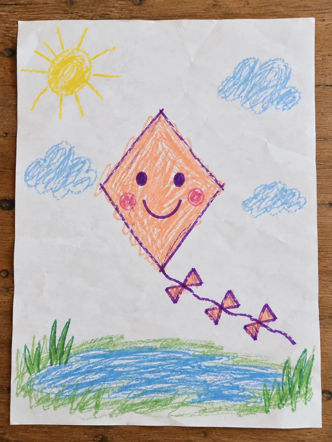 A child's crayon drawing of a smiling kite with three bows above a pond